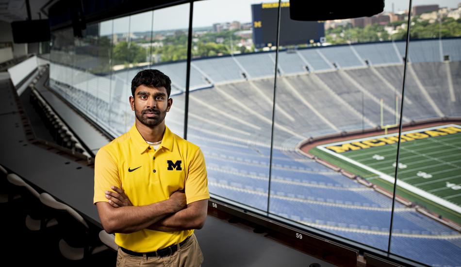 Rohit Mogalayapalli in a box in the big house. The field and bleachers below are empty.  He is wearing a Maize polo. 