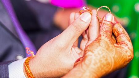 The hands of a bride and groom holding rings. The bride's hand is adorned with intricate henna. 