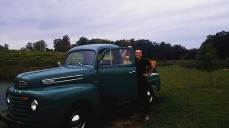 David Hernandez smiles and poses standing next to a vintage green pickup truck parked in a field under a light, cloudy sky.