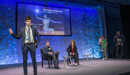 Charlie Logan standing onstage at the MIT Sloan Sports Analytics Conference. Daryl Morey and Jessica Gelman, founders of the conference, sit onstage behind him.