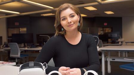 Chloe Preble smiles and sits at a table in the Visualization Study at the James and Ann Duderstadt Center. An Oculus headset and controllers lay on the table in front of her. 