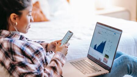 A person lounges with a laptop open on their lap. The screen shows a graph, which they compare against information on their smartphone. 