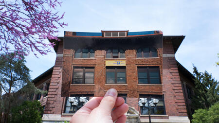 A hand holds up a photo of University of Michigan's West Hall with a School of Information banner hanging from the window in front of the actual West Hall building.