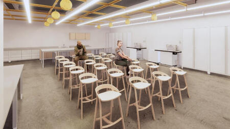 Dozens of modern tall chairs are aligned in a room with flexible furniture and sleek lighting lined across the ceiling, two people sit in the middle