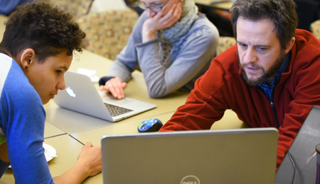 Two students collaborate over a laptop. Another student is working on a laptop in the background.