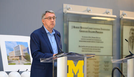 Donor Larry Leinweber speaks at a podium adorned with a Block M. Behind him are commemorative hardhats and a rendering of the Leinweber Computer Science and Information building. He is wearing glasses and a blue blazer.