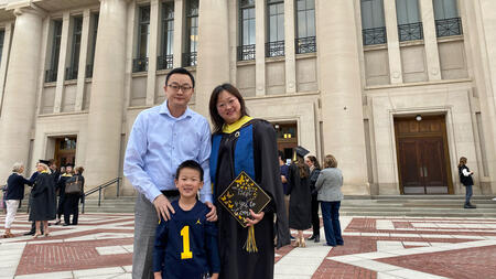 Yao Wei dressed in graduation gown stands outside of an auditorium on University of Michigan campus with her husband and son on her graduation day. She holds a decorated graduation cap.