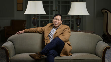 Matthew Bui sitting on a couch wearing a blazer and smiling. 