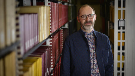 Jesse Johnson wears glasses, a cardigan and stands next to a bookshelf. 