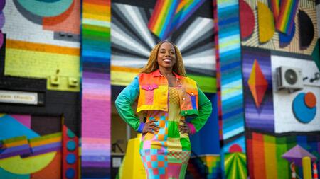 Megan poses in front of a rainbow and prismatic mural in an alleyway while wearing a vibrant dress and jacket of a patchwork of shapes and colors.