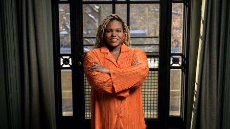 Chelsea Peterson-Salahuddin wearing orange, smiling and standing in front of a window. 