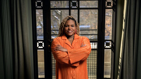 Chelsea Peterson-Salahuddin wearing orange, smiling and standing in front of a window. 