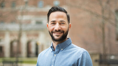 Alain Cohn wearing a button up blue shirt, standing outside, and smiling at the camera. 