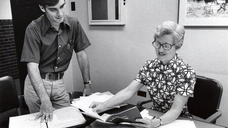 An archival photo of former UMSI professor Constance Rinehart. She is shown sitting at a desk and holding a document. A young man is looking over her shoulder. 