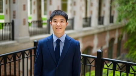 Jiaxin Pei wearing a blue suit and tie, standing outside and smiling at the camera. 