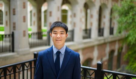 Jiaxin Pei wearing a blue suit and tie, standing outside and smiling at the camera. 