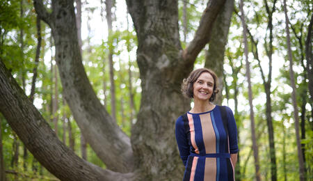 Nicole Ellison in front of a tree on a hiking trail, wearing a blue and pink dress and smiling.  