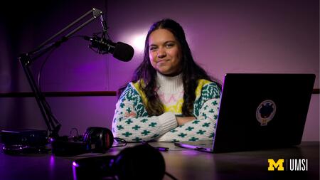 An image of Aayana Anand sitting in front of a podcasting microphone and computer with purple lighting behind her. 