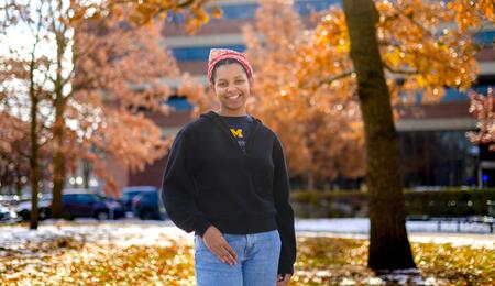 An image of Christine Carethers standing on campus surrounded by fall foliage, wearing a U-M t-shirt and a red bandana