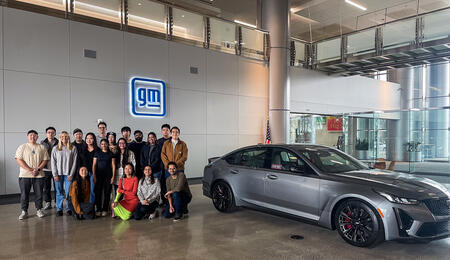 An image of students posing beside a classic Cadillac model in the lobby of General Motors' Cole Building, with an illuminated "gm" sign on the wall behind them