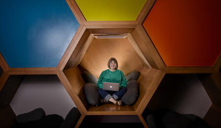 Kristin Fontichiaro sitting cross-legged in colorful cubes at a library, wearing a green sweater and smiling.