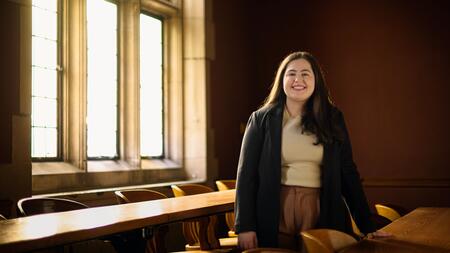 A photo of Julia Kassab posing behind a long wooden desk in an empty Law School classroom