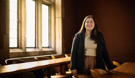 A photo of Julia Kassab posing behind a long wooden desk in an empty Law School classroom