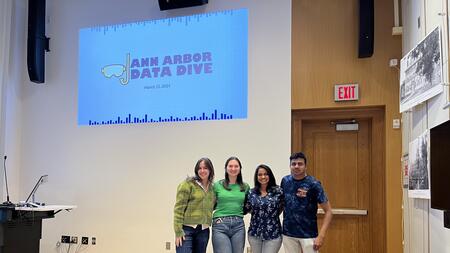 Four students standing together under a projected sign that says "Ann Arbor Data Dive"