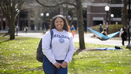 A photo of Ambar Amoros-Gomez standing on the Diag wearing a U-M sweatshirt, with a hammock visible in the background