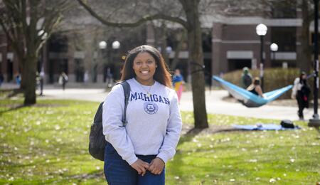 A photo of Ambar Amoros-Gomez standing on the Diag wearing a U-M sweatshirt, with a hammock visible in the background