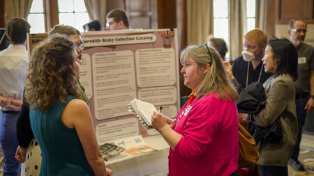 Two students speak with a professor in front of a trifold poster in the Michigan League.