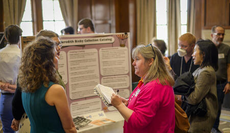 Two students speak with a professor in front of a trifold poster in the Michigan League.