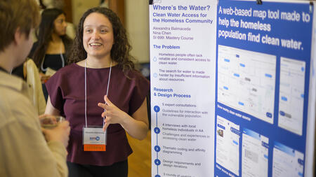 A student stands in front of a trifold poster, chatting animatedly with a guest at Exposition