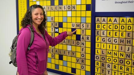 A photo of advisory board member Nikki Roda pointing to "UMSI" on a scrabble board display adhered to a wall
