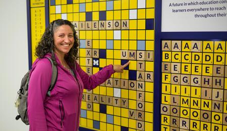 A photo of advisory board member Nikki Roda pointing to "UMSI" on a scrabble board display adhered to a wall