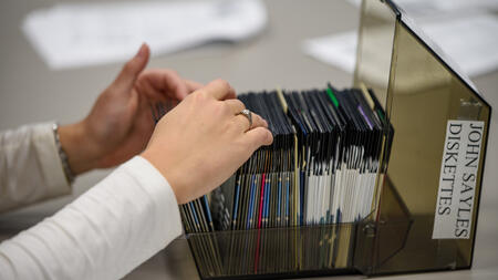 Two hands flipping through a file of diskettes. The label on the file says "John Sayles Diskettes"