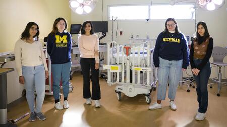 A photo of five students standing in a light-filled operating room. Two of them wear "University of Michigan" sweatshirts.