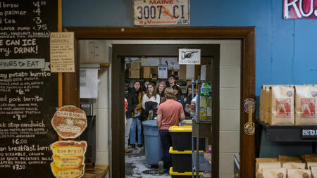 A group of students are visible through a doorway. Framing the door are a café menu and rows of coffee bags.
