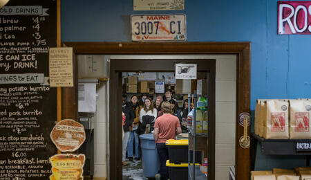 A group of students are visible through a doorway. Framing the door are a café menu and rows of coffee bags.