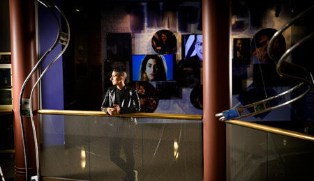 Tam Rayan wears a leather jacket and short hair and leans on a railing at the Arab American National Museum.