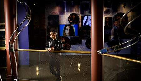 A photo of Tam Rayan posing inside the Arab American National Museum with images projected on a wall behind them