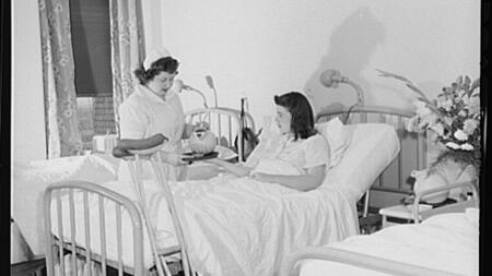 A black and white photo of a teenage nurse's aide pouring a drink for a patient lying in a bed