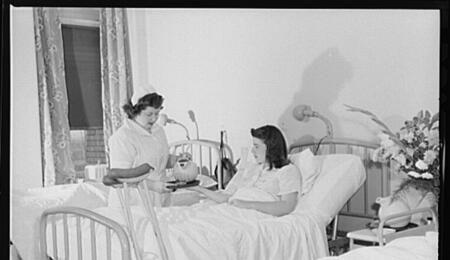 A black and white photo of a teenage nurse's aide pouring a drink for a patient lying in a bed