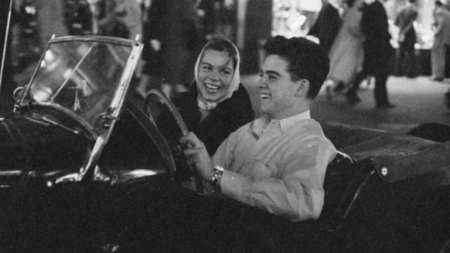 A black and white photo of a teenager driving a classic car while another teen smiles from the passenger seat