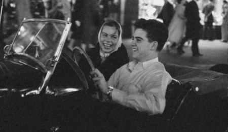 A black and white photo of a teenager driving a classic car while another teen smiles from the passenger seat