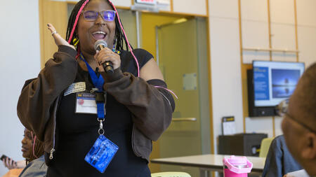 Courtnee Pruitt stands holding a microphone and makes a big gesture with her hand, with a huge smile.
