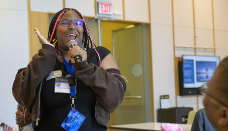 Courtnee Pruitt stands holding a microphone and makes a big gesture with her hand, with a huge smile.