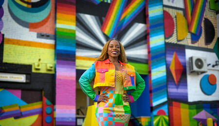 Megan Threats in a colorful jacket and a long skirt standing in front of a multi-colored wall.