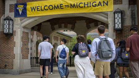 About 10 students walk into an outdoor brick passageway that is decorated with a maize and blue banner that reads "Welcome to Michigan." Two of the students wear UMSI-branded backpacks.