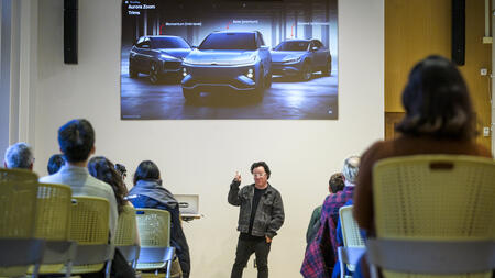A person giving a presentation to a room full of people. A photo of three cars is projected on a screen above him.